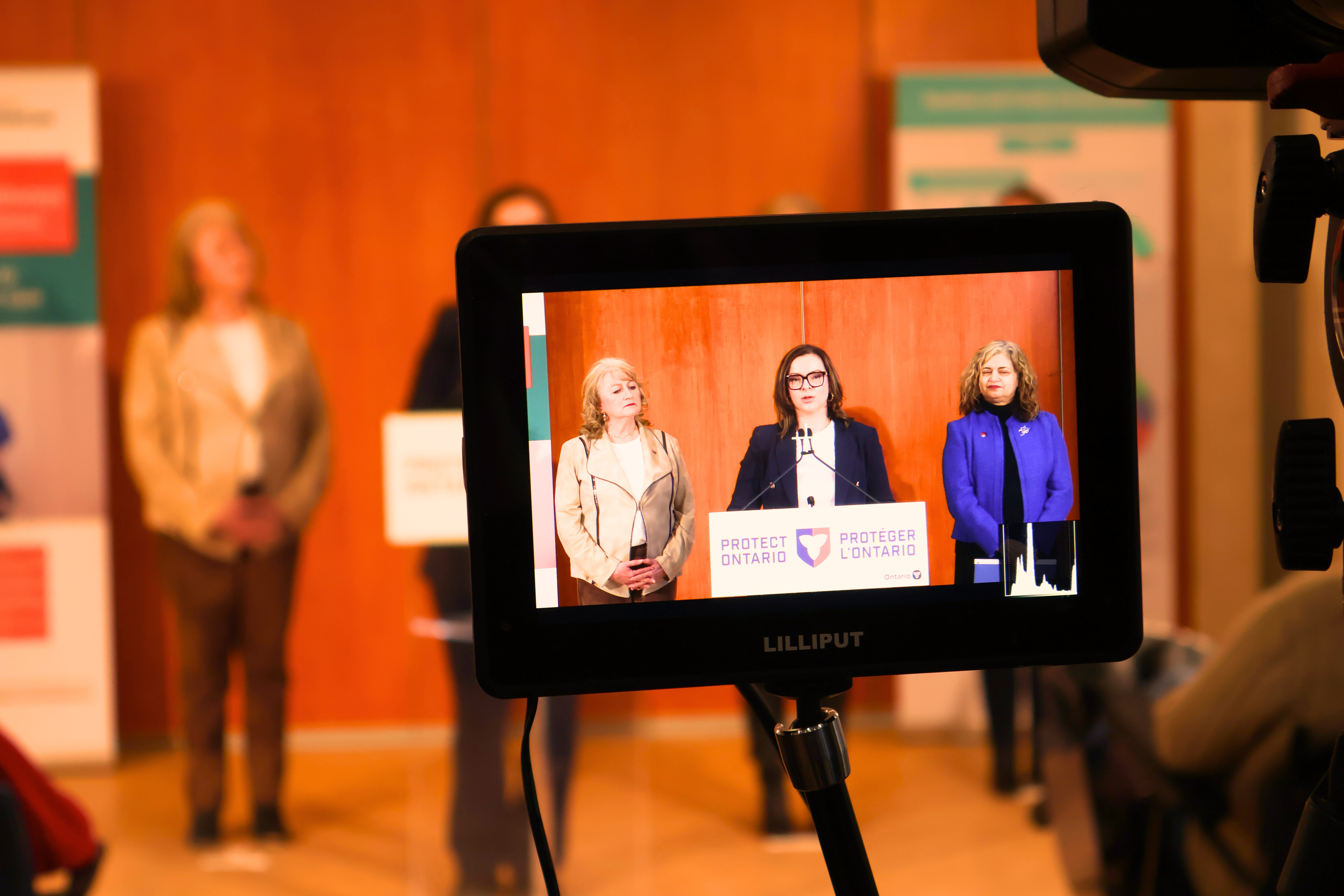A camera monitor displays three speakers standing at a podium labeled Protecting Ontario during an announcement related to the $288,988 Improving Dementia Care Program Grant, with the event setting and additional attendees visible in the background.