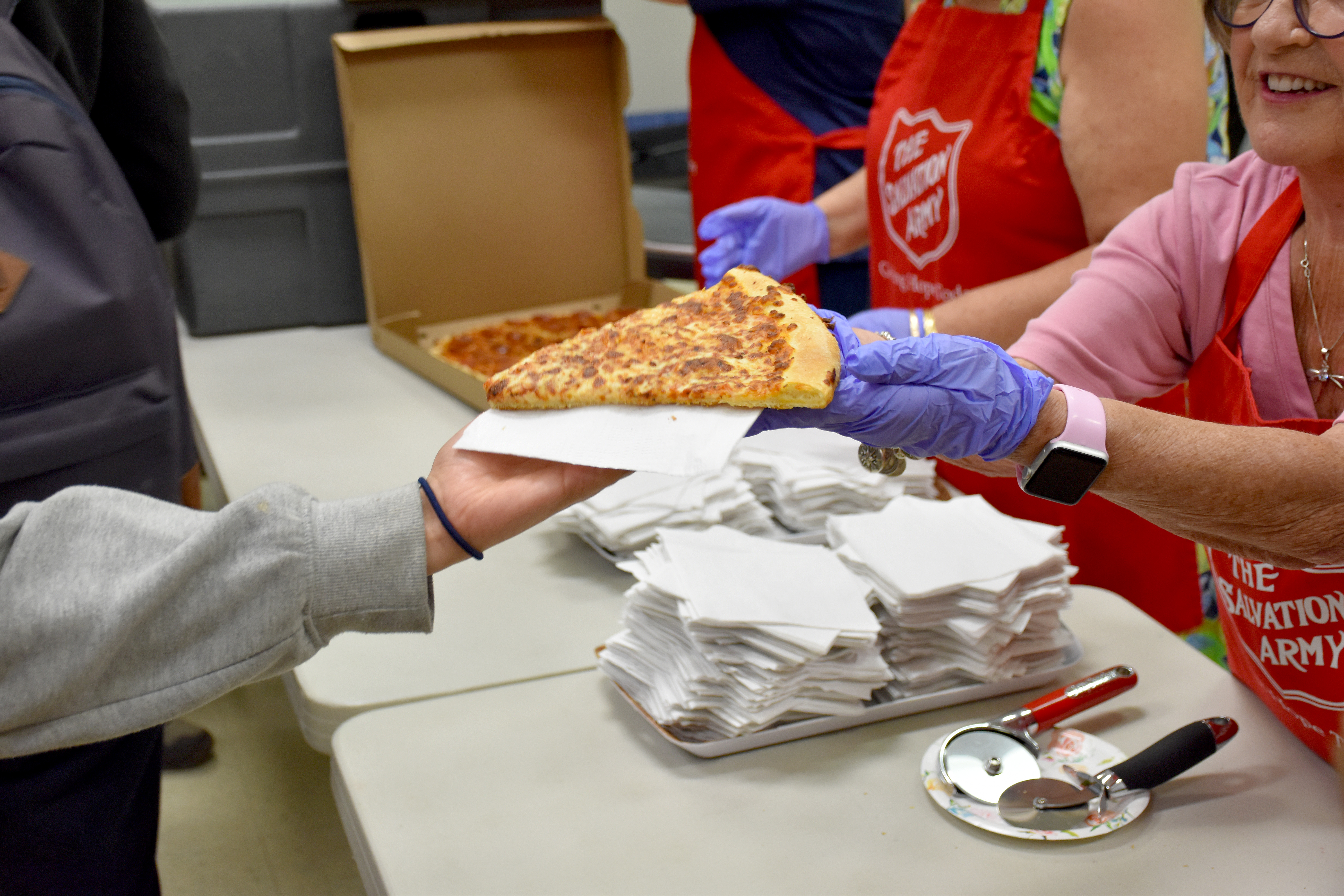The Salvation Army volunteer wearing gloves hands a slice of pizza to a resident across a table.