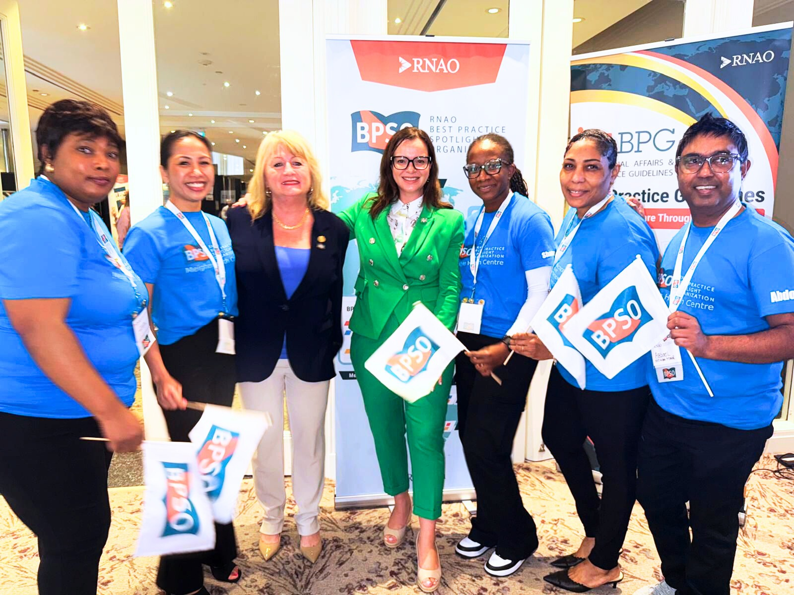 A group of people wearing blue event shirts stand together holding BPSO flags in front of banners for the Best Practice Spotlight Organization (BPSO) Designation event, posing beside two formally dressed attendees.
